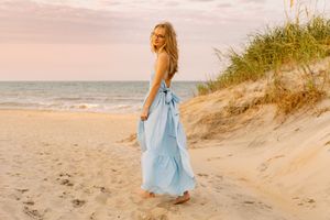 A young blonde woman in a light blue maxi dress walks barefoot on a sandy beach at sunset.