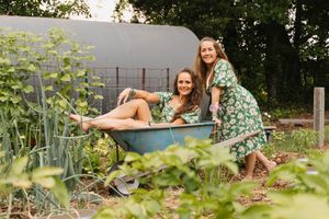 Two women in green floral dresses laugh together while sitting in a blue garden wheelbarrow outdoors.