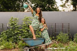 Two women in matching green floral dresses pose playfully in a garden wheelbarrow, one holding a watering can.