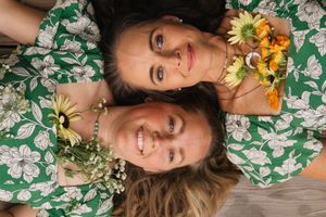 Two women in matching green floral dresses lie head-to-head holding sunflowers and wildflowers.