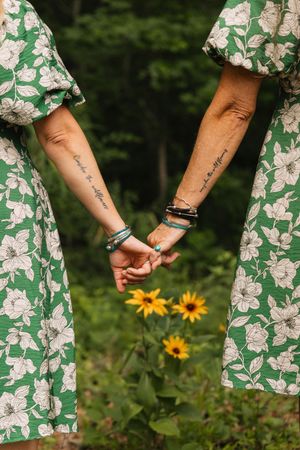 Two women in matching green floral dresses hold hands showing matching script tattoos and turquoise bracelets with yellow wildflowers.