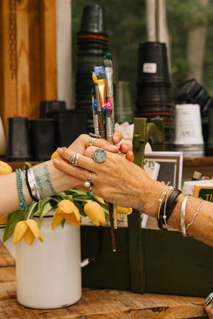 Two hands adorned with rings and bracelets hold paintbrushes near yellow tulips on a wooden table.