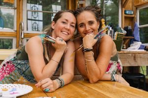 Two smiling women pose with paintbrushes at a wooden table during an art activity outdoors.