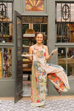 Woman in floral apron holding paintbrush and palette stands in doorway of charming boutique shop.
