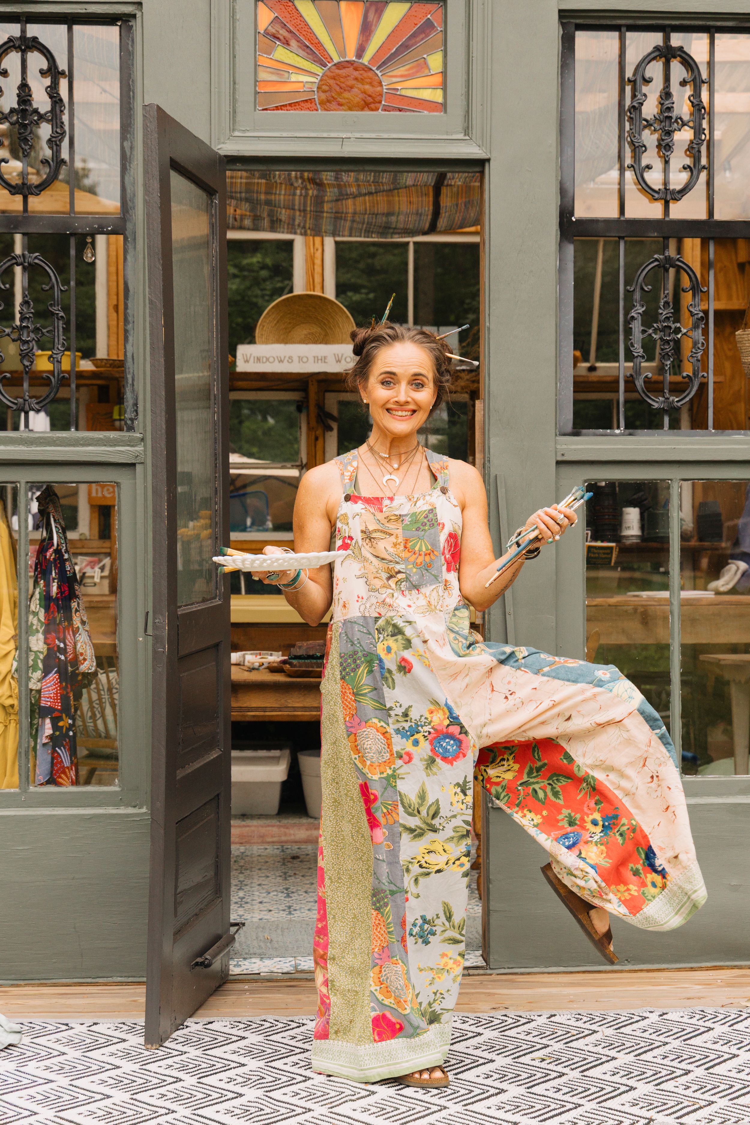 Woman in floral apron holding paintbrush and palette stands in doorway of charming boutique shop.
