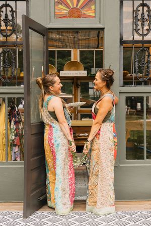 Two women in colorful patchwork maxi dresses smile at each other outside a boutique shop entrance.
