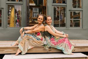 Two women in colorful boho maxi skirts sit back-to-back smiling on wooden steps outside a boutique shop.