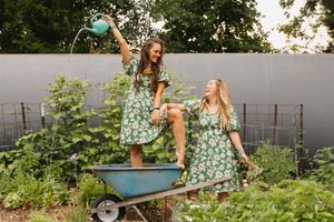 Two women in green floral dresses play in a garden, one watering the other sitting in a wheelbarrow.