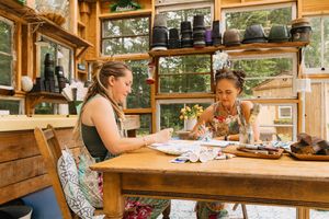 Two women painting at a rustic wooden table in a sunlit greenhouse studio with shelves of pots.