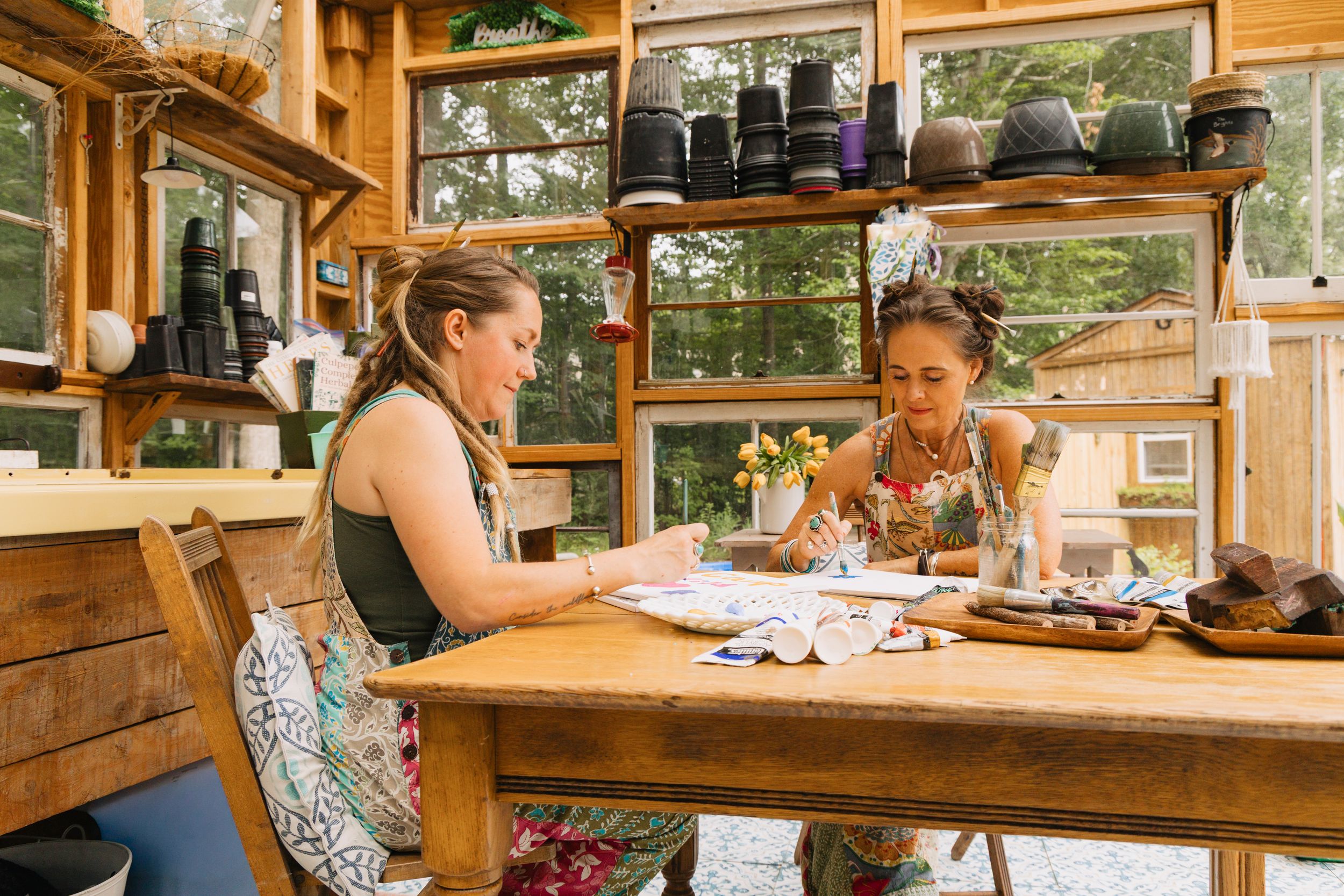 Two women painting at a rustic wooden table in a sunlit greenhouse studio with shelves of pots.