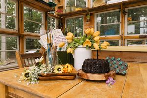 Rustic wooden table with yellow tulips, bundt cake, and floral decorations in a sunlit greenhouse.