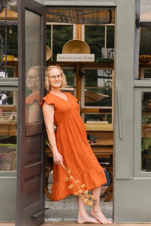 A barefoot woman in an orange midi dress stands in an open doorway of a charming boutique shop.