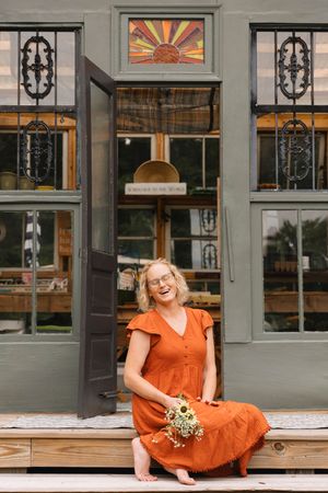 Cheerful blonde woman in orange dress sits on steps of a charming green boutique storefront.