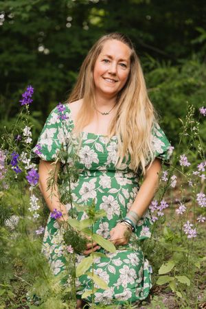 Smiling blonde woman in green floral dress holds wildflowers in a lush garden setting.