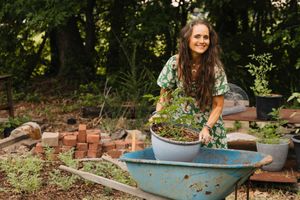 Woman smiling while gardening with a wheelbarrow filled with plants outdoors.