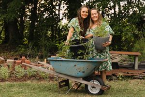 Two women in matching green floral dresses smile holding plants beside a blue wheelbarrow in a garden.