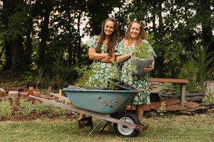 Two women in matching green floral dresses pose with a wheelbarrow full of plants outdoors.