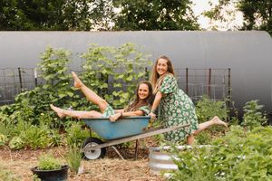 Two women in green floral dresses laugh as one sits in a wheelbarrow in a lush garden.