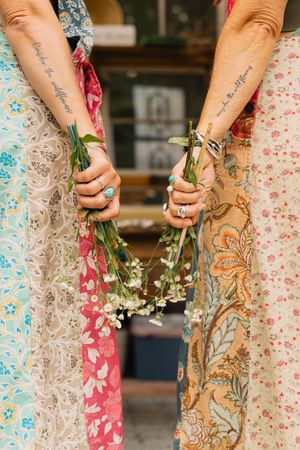 Two women in colorful boho floral skirts hold white wildflowers together displaying matching forearm script tattoos.
