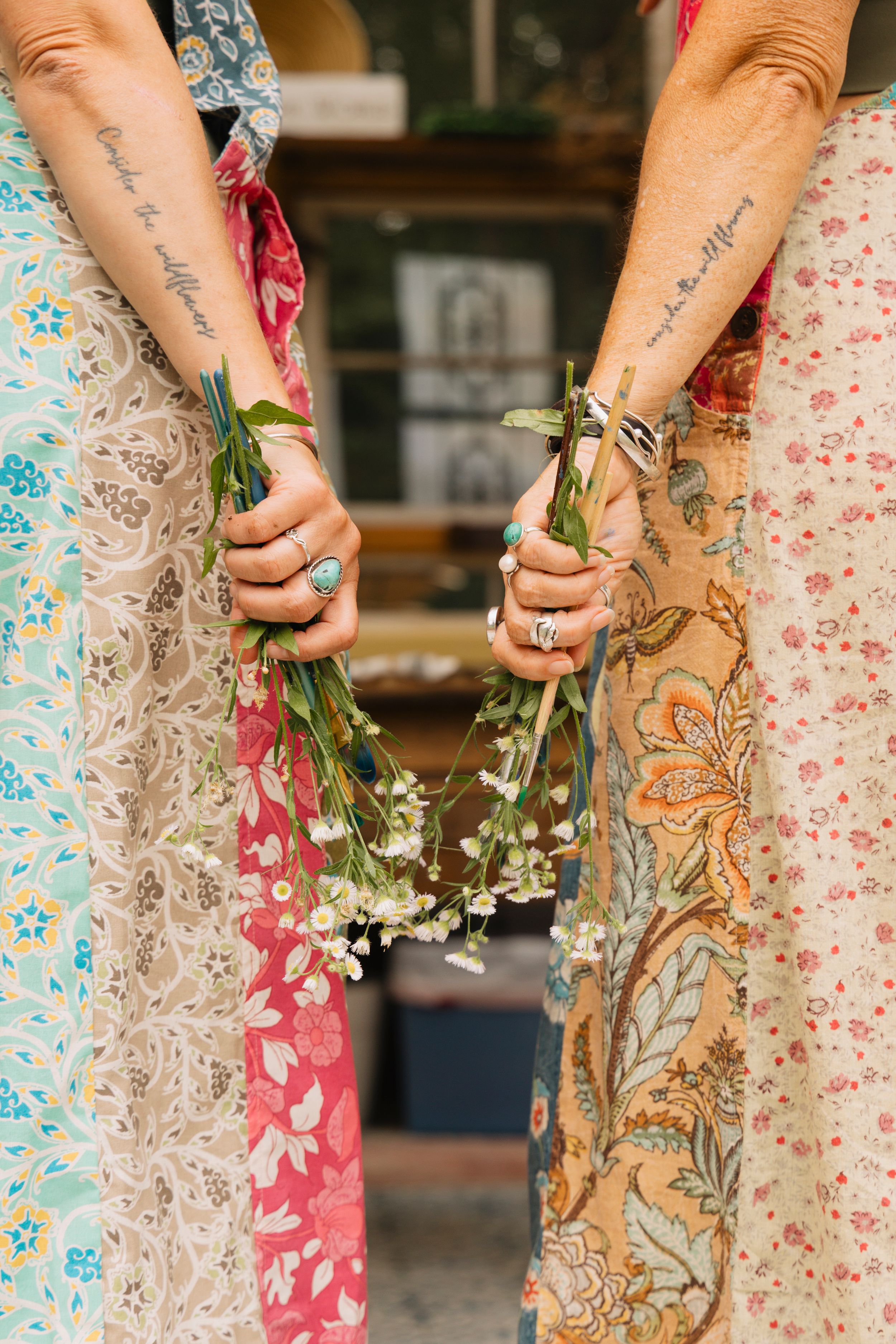 Two women in colorful boho floral skirts hold white wildflowers together displaying matching forearm script tattoos.