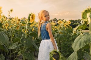 Young girl in denim top and white skirt standing among tall sunflowers in a golden sunset field.