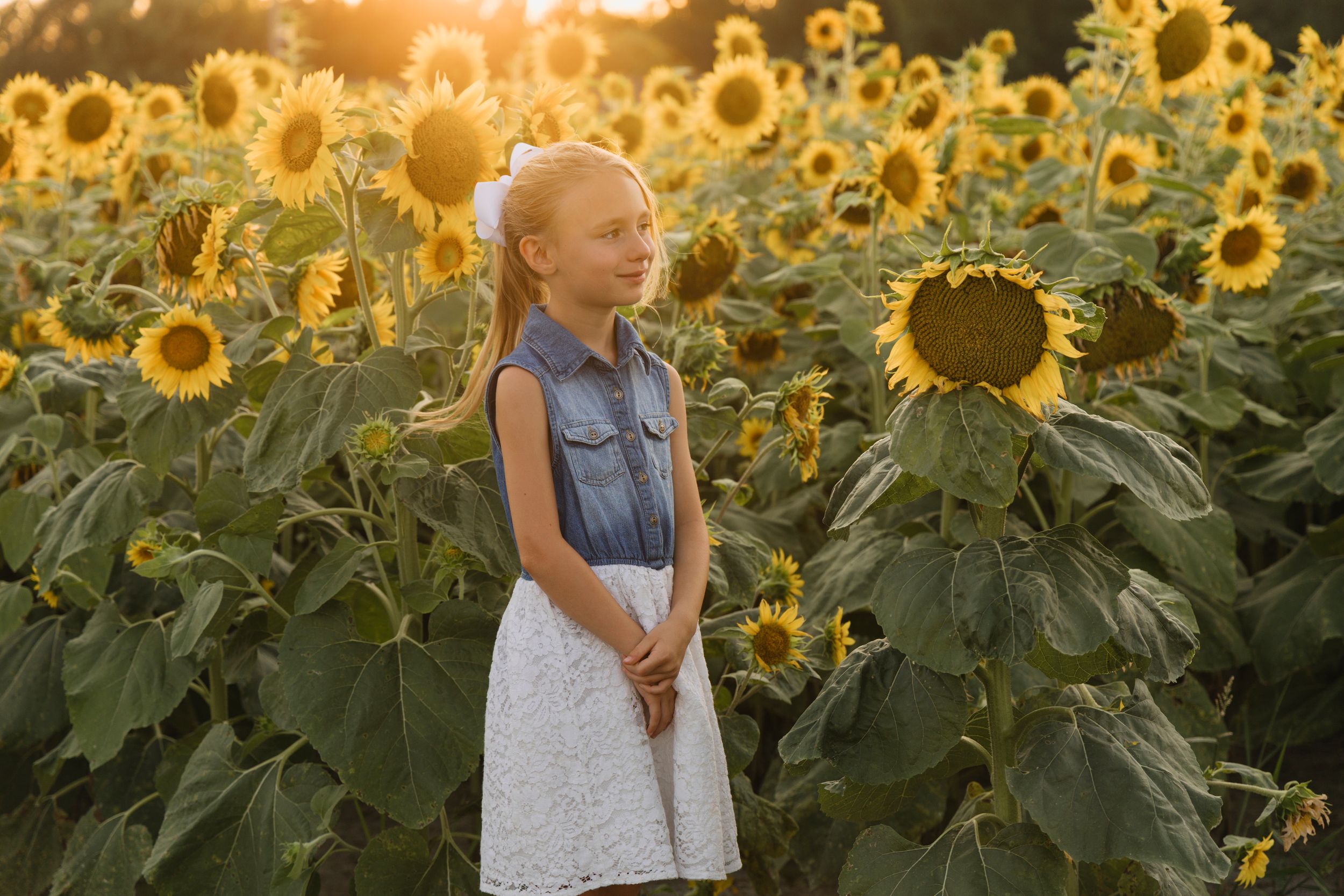 Young girl in white skirt and denim vest standing in a vibrant sunflower field during golden hour sunset.