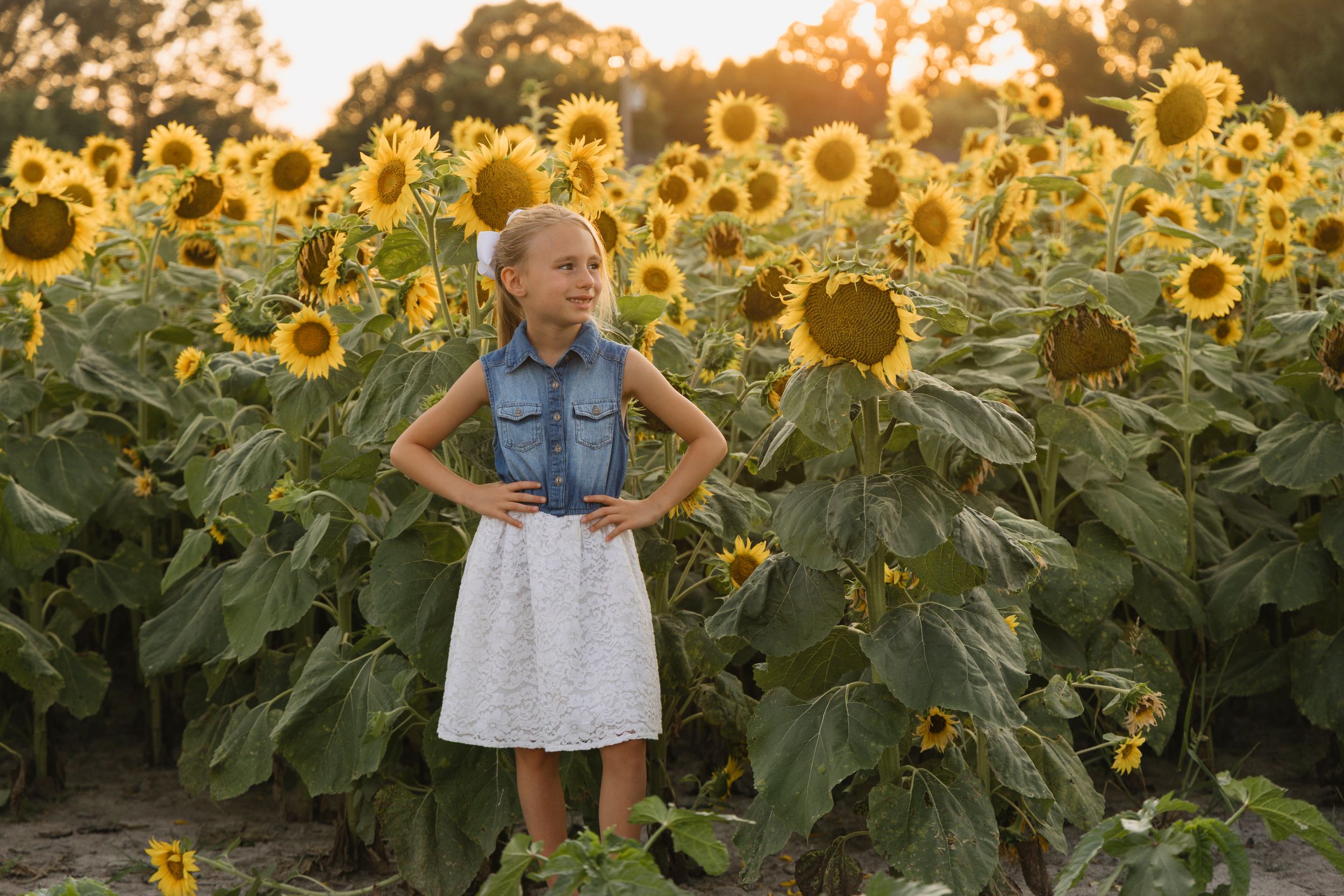 Girl in denim vest and white skirt stands confidently in a vibrant sunflower field during golden hour.