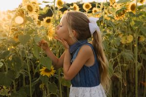 Young girl with long hair and white bow blowing a kiss to sunflowers in a golden sunlit field.