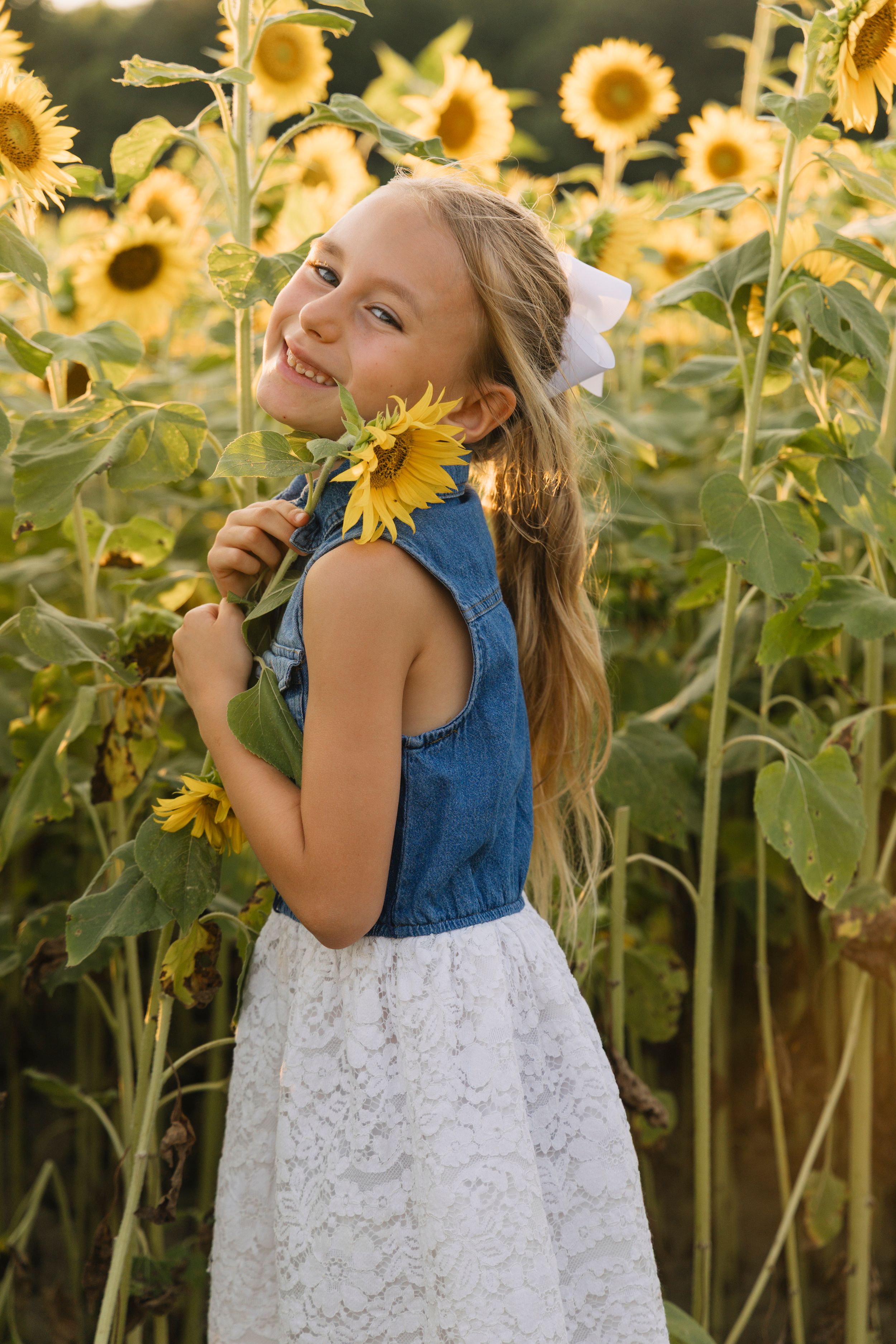 Smiling girl in denim top and white lace skirt holding a sunflower in a sunny sunflower field.
