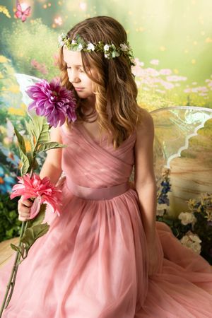 Young girl in a pink tulle dress and floral crown holds pink dahlias, with sheer fairy wings in a dreamy garden backdrop.