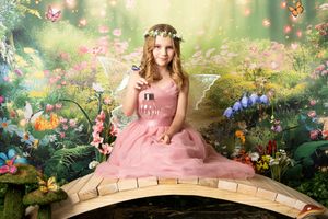 Girl in a pink tulle dress and floral crown with fairy wings sits on a wooden bridge in an enchanted garden with butterflies and flowers.