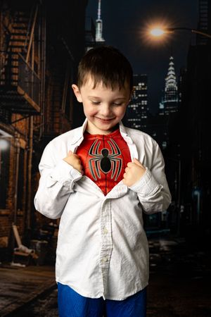 Young boy smiling, revealing Spider-Man costume under white shirt, posed against NYC cityscape backdrop.