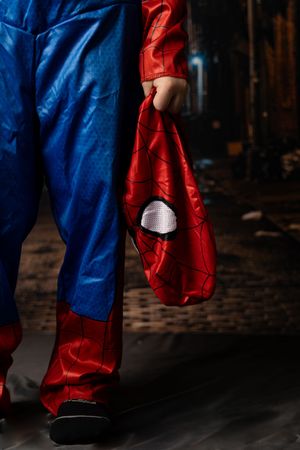 Person in Spider-Man costume holding the hero mask while wearing blue and red superhero suit in a dark alley.