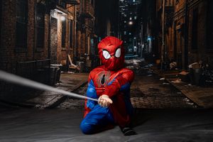 Child in Spider-Man costume kneeling in dark alley at night, holding a web prop in urban setting.