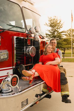 Couple posing on a red fire truck, woman in orange dress seated while man in firefighter gear smiles beside her.