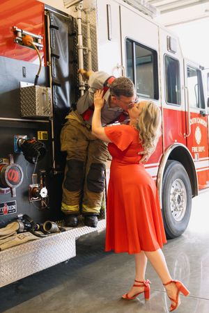 A woman in a red dress kisses a firefighter in uniform beside a red fire truck in a station.