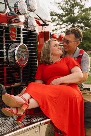 A couple poses romantically on a red fire truck, woman in red dress and heels sitting on mans lap outdoors.