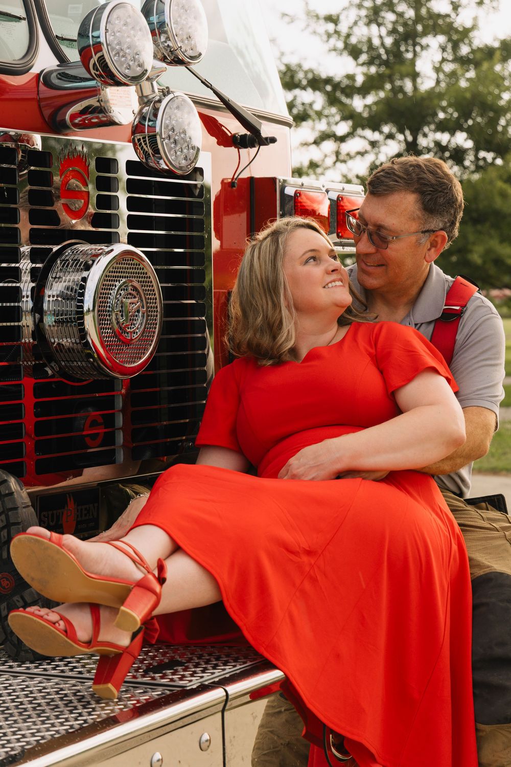 A couple poses romantically on a red fire truck, woman in red dress and heels sitting on mans lap outdoors.