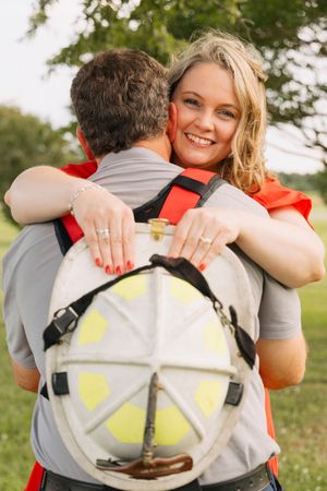 Smiling blonde woman hugging a firefighter in uniform outdoors, showing off an engagement ring with red nails.