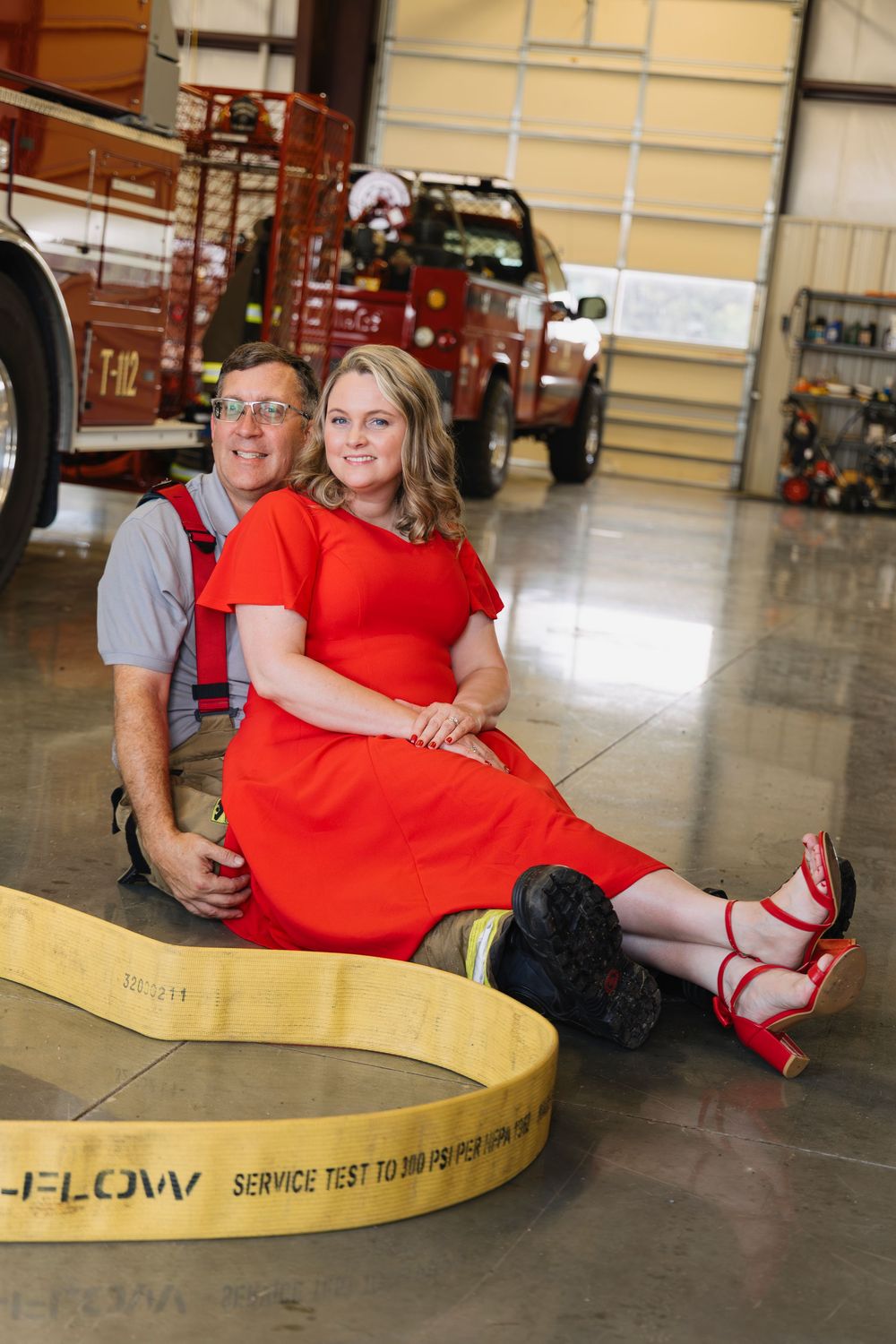 Couple posing inside a fire station with red fire trucks, woman in red dress seated on a fire hose, man in firefighter suspenders.
