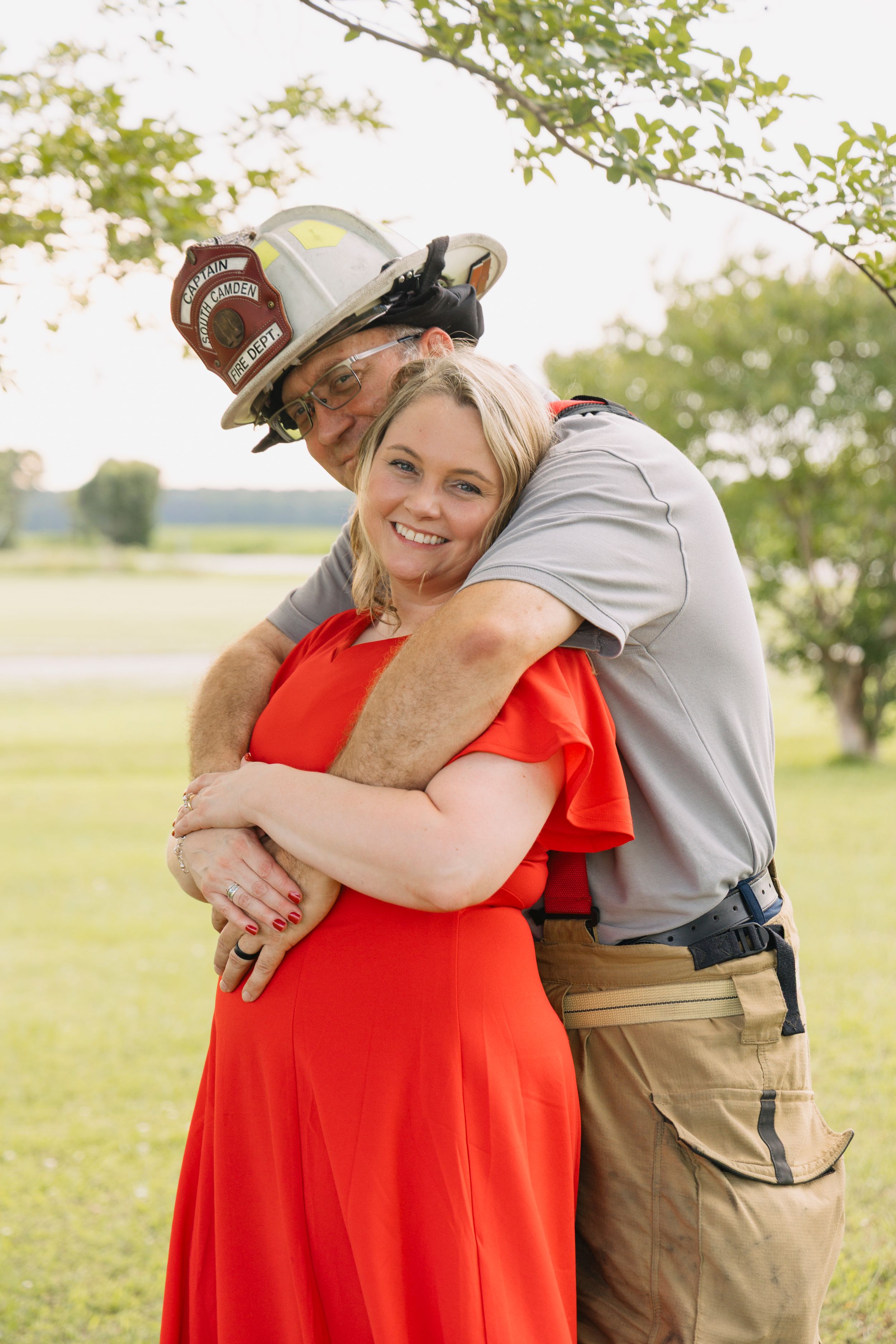 A firefighter in helmet hugs a smiling woman in a red dress outdoors in a green field.