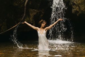 Woman in white dress splashing in a waterfall pool with arms raised in a lush rocky setting.
