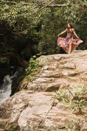 Woman in floral skirt dancing on rocky cliffs beside a waterfall surrounded by lush green forest trees.