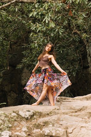 Woman in colorful flowing skirt and brown top posing on rocky terrain surrounded by lush green foliage.