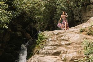 Woman in colorful maxi dress standing on rocky steps near a cascading waterfall in lush woods.
