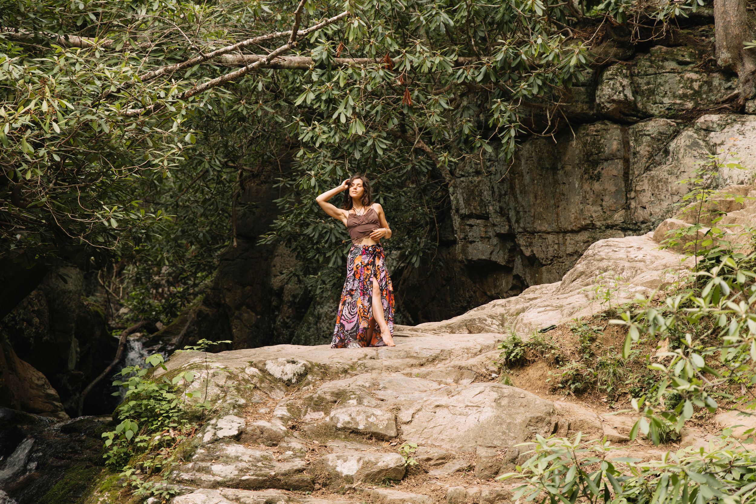 Woman in floral maxi skirt poses on rocky outcrop surrounded by lush green trees and stone cliff backdrop.