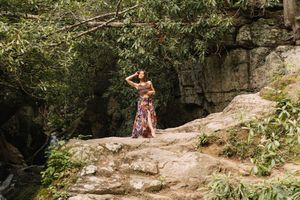 Woman in floral maxi skirt poses on rocky outcrop surrounded by lush green trees and stone cliff backdrop.