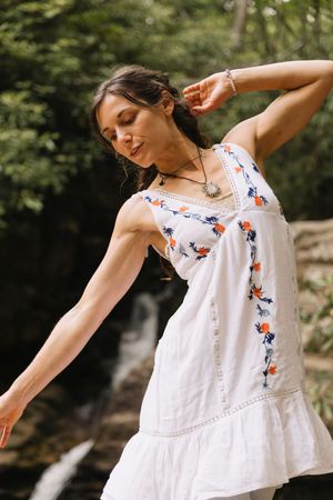 Woman in white floral embroidered sundress dancing gracefully in front of a lush green forest waterfall.