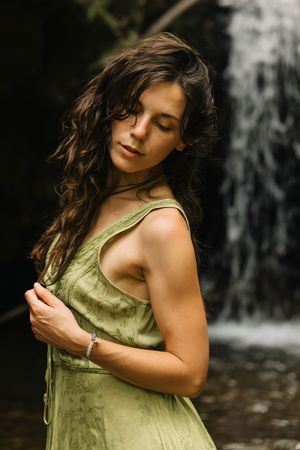 Woman in olive green dress with eyes closed standing before a cascading waterfall in nature.