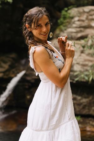 Smiling woman in white embroidered dress holding a daisy near a waterfall in a lush green forest.